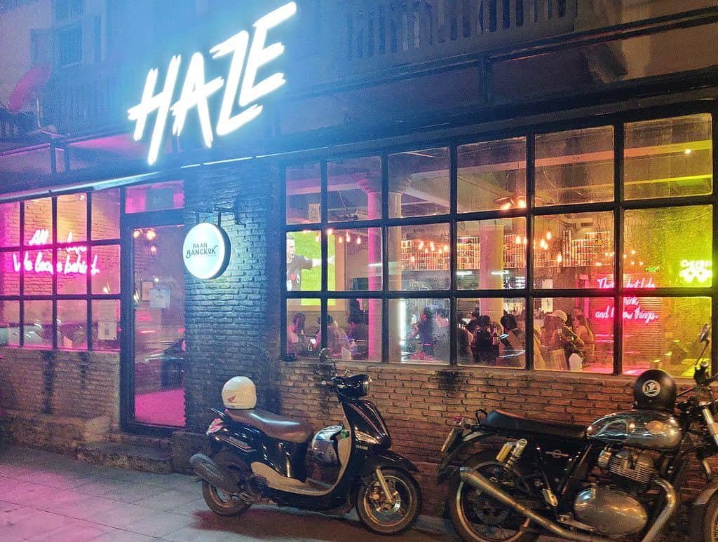 View of Haze bar from outside at night in Bangkok Thailand. Large open glass windows show bright neon lights inside the craft beer bar. 