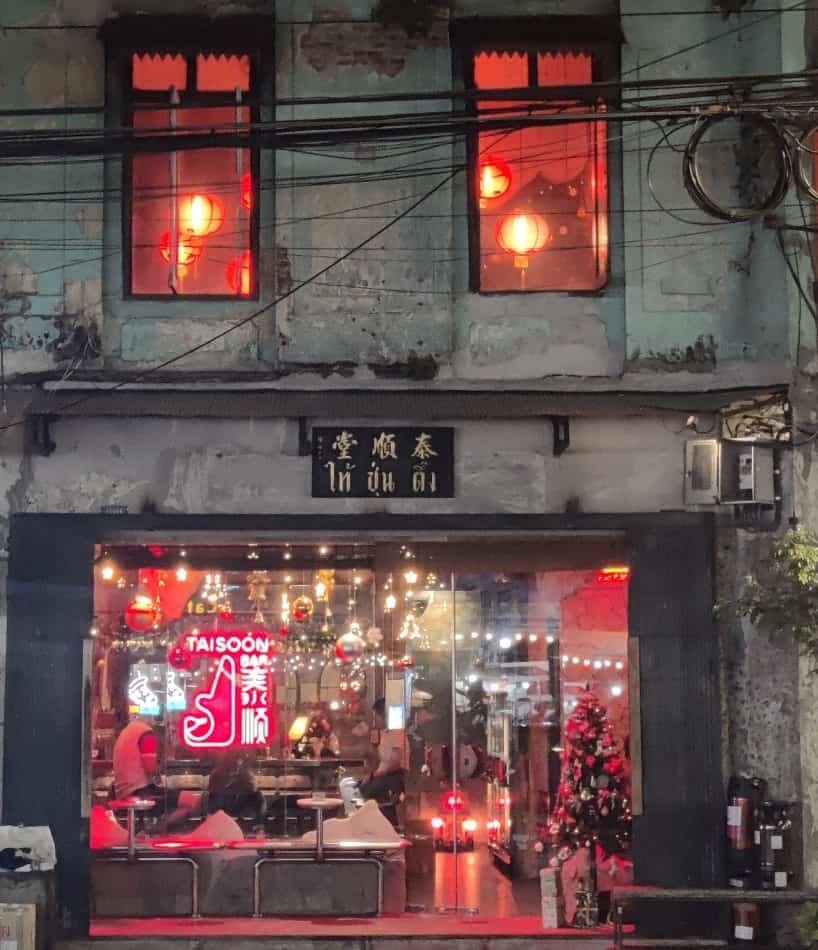 Outside view of Tai Soon Bar in the evening. Red lanterns showing through the upstairs windows, and lights and decoration showing through the glass wall of the bar's first floor. 