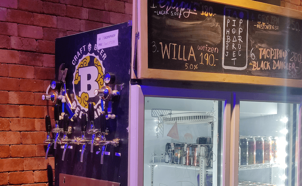 Six beer taps on the wall of the fridge against a brick wall. Chalk board lists craft beer available in Taopiphop bar project Bangkok.