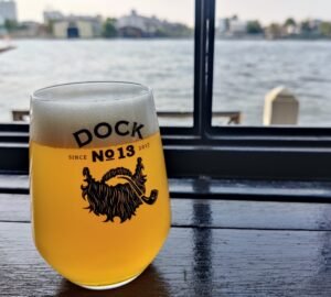 Local Thai craft beer in a Dock No.13 branded glass - messy beard and pipe under the logo. Glass on a dark wood table with the river in the background as sunlight comes from behind