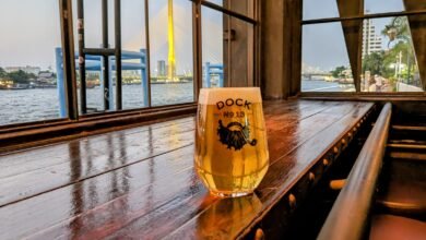Glass of beer on a wooden table with water in the background and the sunset on a bridge. At Sheepshank Public house or Dock No. 13 in Bangkok Thailand.